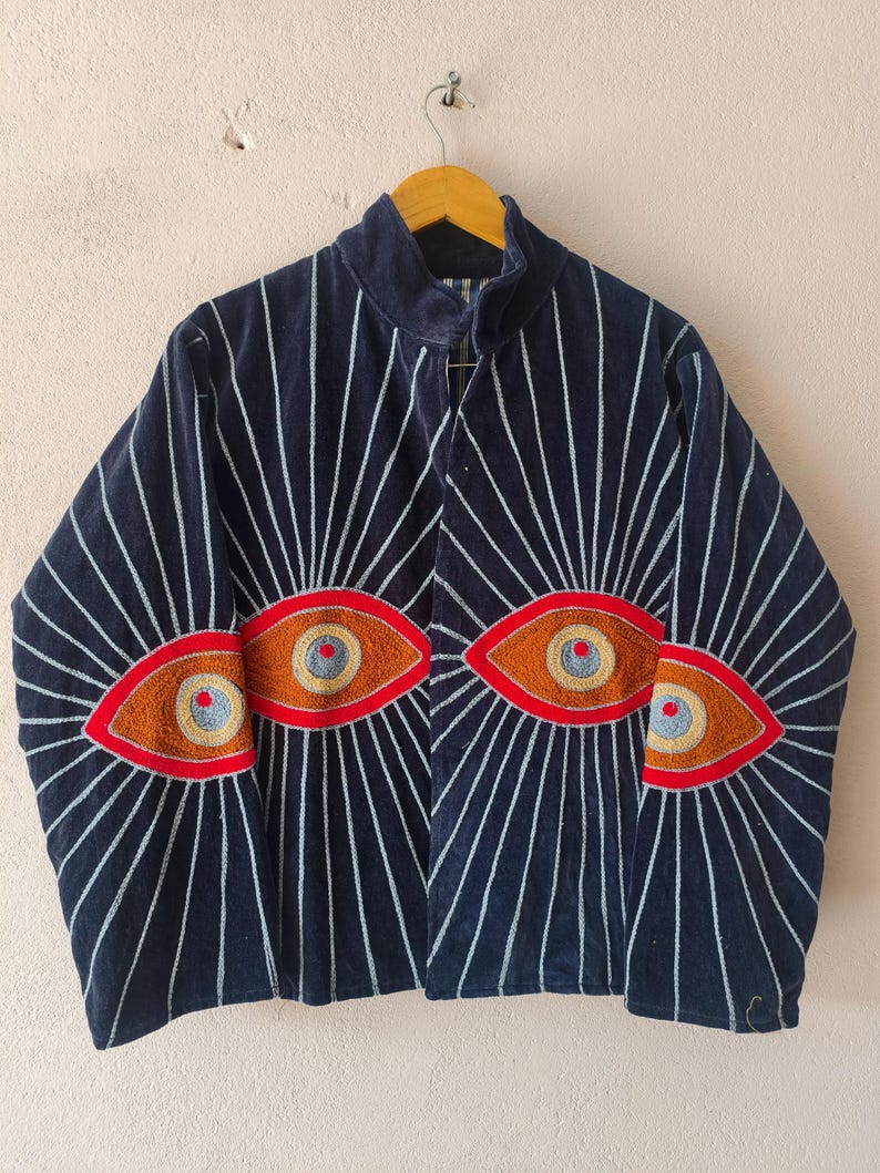 Puede incluir: Chaqueta azul marino con l&iacute;neas verticales blancas que irradian desde el centro. La chaqueta presenta tres dise&ntilde;os de ojos bordados en tonos naranja, rojo y azul. La chaqueta est&aacute; colgada en una percha de madera.