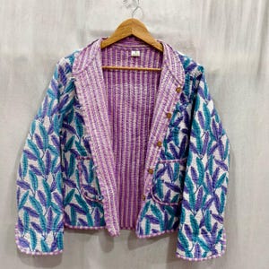 May include: A quilted jacket with a leaf pattern in shades of teal, purple, and white. The jacket has a purple and white striped interior and a notched collar. It features wooden buttons and two front pockets. The jacket is hanging on a wooden hanger.