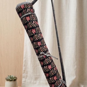 May include: A black yoga mat bag with a floral pattern in red and green. The bag has a striped top and bottom, and a black and white striped strap. The bag is hanging on a gold hook.