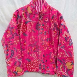 以下が含まれることがあります： 鮮やかなピンク色のジャケット。花柄のデザインで、ハイネックとフルレングスのジッパーが付いています。花柄には赤、青、黄色のアクセントがあります。柔らかくビロードのような素材で作られています。