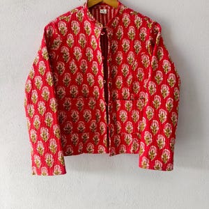 Könnte beinhalten: Eine gesteppte Jacke in leuchtendem Rot mit einem sich wiederholenden Blumenmuster in Rosa- und Grüntönen. Die Jacke hat einen Stehkragen, lange Ärmel, eine Knopfleiste auf der Vorderseite und zwei aufgesetzte Taschen, hängend an einem Kleiderbügel.