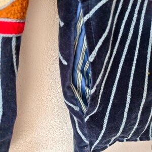 Puede incluir: Primer plano de una chaqueta azul marino con rayas verticales azul claro. La chaqueta tiene un bolsillo con forro a rayas azul y blanco. La manga presenta un dise&ntilde;o de ganchillo colorido con colores rojo, naranja y crema.