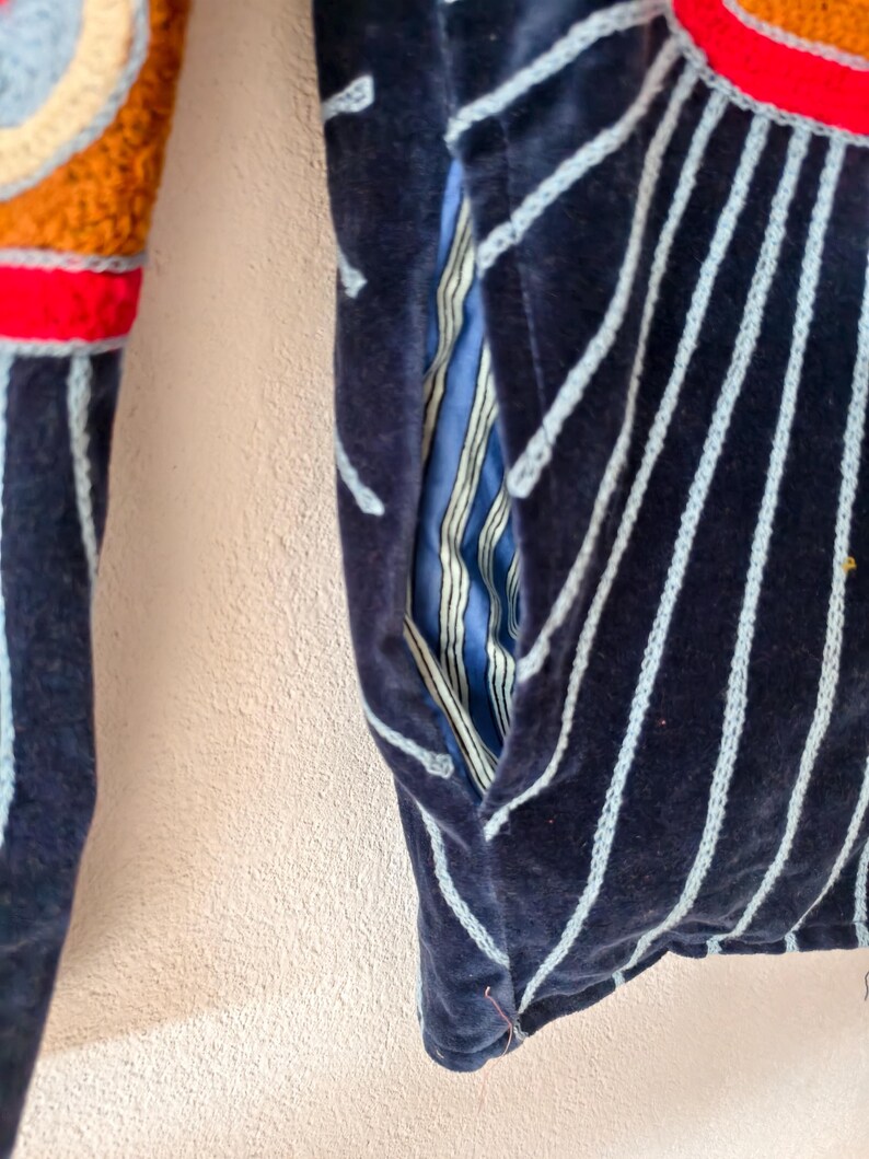 Puede incluir: Primer plano de una chaqueta azul marino con rayas verticales azul claro. La chaqueta tiene un bolsillo con forro a rayas azul y blanco. La manga presenta un dise&ntilde;o de ganchillo colorido con colores rojo, naranja y crema.