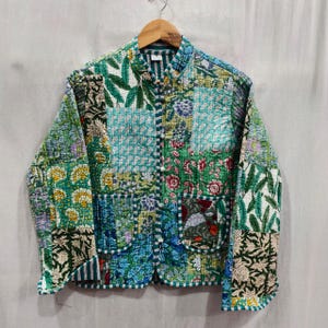 May include: A patchwork jacket with a mandarin collar and button closure. The jacket features a variety of floral and leaf patterns in shades of green, blue, and white. Two pockets are visible on the front. The jacket is hanging on a wooden hanger.