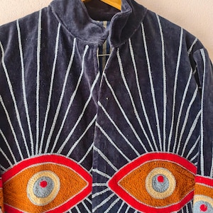 Puede incluir: Chaqueta de terciopelo azul marino con rayas verticales blancas y dise&ntilde;os de ojos bordados. La chaqueta tiene un cuello alto y est&aacute; adornada con grandes motivos de ojos coloridos en tonos rojos, naranjas y grises, creando un look &uacute;nico y art&iacute;stico.