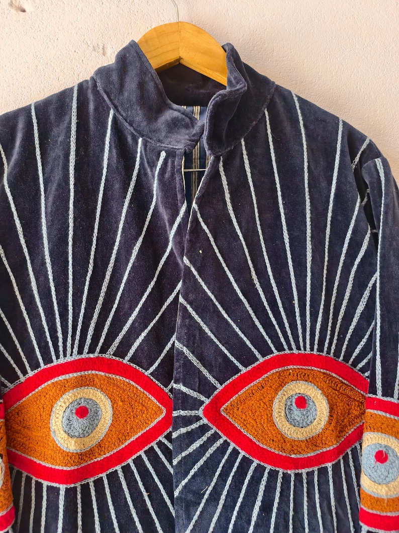 Puede incluir: Chaqueta de terciopelo azul marino con rayas verticales blancas y dise&ntilde;os de ojos bordados. La chaqueta tiene un cuello alto y est&aacute; adornada con grandes motivos de ojos coloridos en tonos rojos, naranjas y grises, creando un look &uacute;nico y art&iacute;stico.