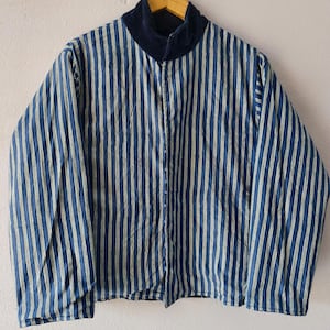 Puede incluir: Una chaqueta a rayas azules y blancas con cuello de terciopelo azul marino cuelga de una percha de madera. La chaqueta tiene una cremallera completa y mangas largas. Las rayas verticales est&aacute;n espaciadas uniformemente.