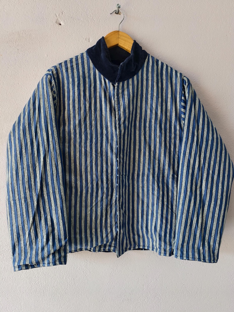 Puede incluir: Una chaqueta a rayas azules y blancas con cuello de terciopelo azul marino cuelga de una percha de madera. La chaqueta tiene una cremallera completa y mangas largas. Las rayas verticales est&aacute;n espaciadas uniformemente.