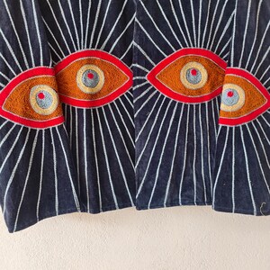 Puede incluir: Chaqueta de terciopelo azul marino oscuro con dise&ntilde;os de ojos bordados. La chaqueta presenta un patr&oacute;n de l&iacute;neas blancas que irradian de los dise&ntilde;os de ojos. Los ojos est&aacute;n delineados en rojo, con detalles marrones, amarillos y azules. Es una prenda &uacute;nica.