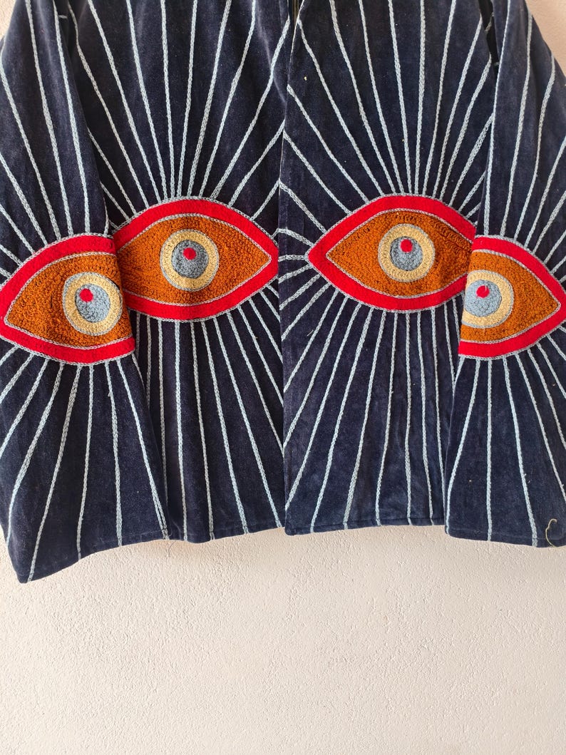 Puede incluir: Chaqueta de terciopelo azul marino oscuro con dise&ntilde;os de ojos bordados. La chaqueta presenta un patr&oacute;n de l&iacute;neas blancas que irradian de los dise&ntilde;os de ojos. Los ojos est&aacute;n delineados en rojo, con detalles marrones, amarillos y azules. Es una prenda &uacute;nica.