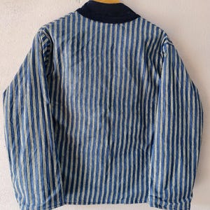 Puede incluir: Chaqueta a rayas azules y blancas con cuello azul marino, colgada de una percha de madera. La chaqueta tiene rayas verticales y mangas largas. La chaqueta est&aacute; hecha de un material suave y ligero.