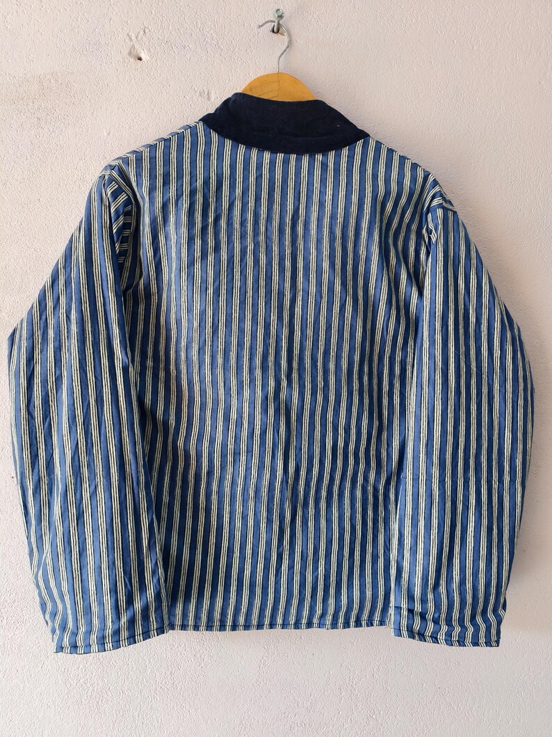 Puede incluir: Chaqueta a rayas azules y blancas con cuello azul marino, colgada de una percha de madera. La chaqueta tiene rayas verticales y mangas largas. La chaqueta est&aacute; hecha de un material suave y ligero.