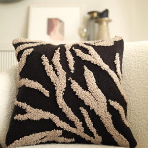 May include: Square black decorative pillow with a beige, textured, abstract pattern. The pillow is on a white couch. The design resembles a zebra print. The pillow is a home decor item.
