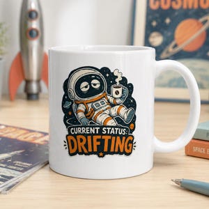 Könnte beinhalten: Weiße Keramik-Tasse mit einem Cartoon-Astronauten-Design. Der Astronaut, in einem Raumanzug, hält eine Kaffeetasse, mit dem Text "CURRENT STATUS: DRIFTING" in Orange und Weiß, vor einem Sternenhintergrund.