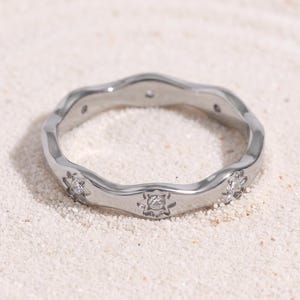 May include: A silver-toned metal ring featuring a wavy, undulating band design. The surface of the ring is accented with small, star-shaped engravings, each containing a single, clear, sparkling gemstone set in the centre of the star motif.