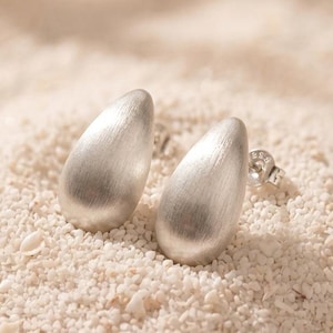 Peut inclure: Une paire de boucles d'oreilles à clous argentées présentant un design en forme de goutte lisse avec une finition mate brossée. Chaque boucle d'oreille possède une fermeture à tige et un poinçon visible sur la pièce métallique près de la base.