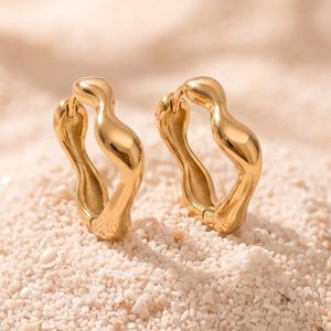 May include: A pair of gold hoop earrings with a distinct wavy, organic silhouette. The earrings feature a polished, reflective surface and a hinged closure mechanism, showcasing a fluid, irregular shape that contrasts with traditional circular hoops.