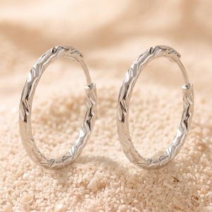 May include: A pair of silver hoop earrings featuring a textured, faceted surface that catches the light. The earrings are circular in shape with a thin wire post closure, showcasing a polished metallic finish and a distinct, repeating geometric pattern.