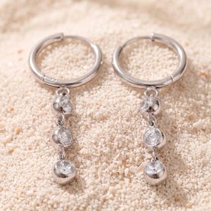 May include: A pair of silver-toned hoop earrings, each featuring a vertical drop attachment consisting of three round, clear, faceted stones set in metal bezels that hang from the bottom of the circular hoop frame.