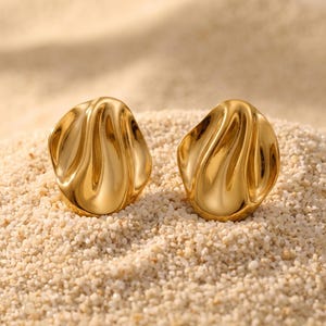 May include: A pair of gold-toned stud earrings featuring an irregular, organic, and wavy shape with a smooth, reflective surface. The earrings have a rounded, molten-like appearance with deep, flowing indentations across the front of each piece.
