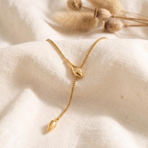 Op de afbeelding: Een goudkleurige lariat-ketting met een slangvormig middendeel met twee kleine heldere steentjes als ogen. De ketting heeft een fijn box-chain ontwerp dat naar beneden loopt en eindigt in een gladde, taps toelopende goudkleurige metalen hanger.