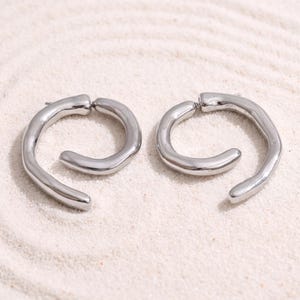Pode incluir: Um par de brincos de argola em tom prateado com acabamento liso e polido. Cada brinco apresenta uma forma circular aberta e irregular com uma banda de metal espessa e arredondada que se curva para dentro, criando uma silhueta espiral.