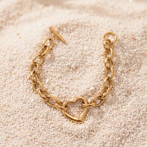 May include: A gold-toned chain bracelet featuring a series of oval links, a central open heart-shaped charm, and a toggle clasp closure consisting of a circular ring and a T-bar component, all crafted with a polished metallic finish.