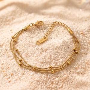 May include: A gold-toned multi-strand bracelet featuring three delicate snake chains adorned with small, evenly spaced round beads. The bracelet includes a lobster claw clasp, an adjustable extender chain, and a small rectangular tag marked S.steel.