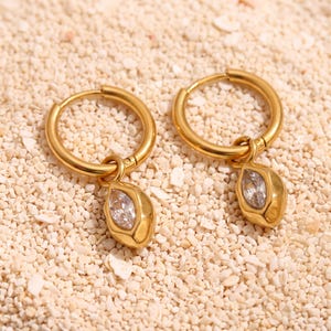 May include: A pair of gold-toned hoop earrings, each featuring a dangling charm with a marquise-cut clear gemstone set in a smooth, polished gold-coloured metal casing. The earrings have a circular hoop design and a secure closure mechanism.