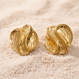 May include: A pair of gold-toned earrings featuring a rounded, abstract shape with a textured, rippled surface. The design consists of smooth, curved edges that wrap around a central, indented section, creating a reflective, metallic finish.