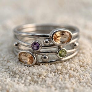 May include: A silver ring with multiple bands, each adorned with oval and round gemstones in shades of peach, purple, and green. The ring is set against a backdrop of small, white pebbles, creating a contrast of textures and colors.