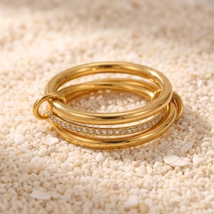 May include: A gold-toned ring featuring three interconnected bands. Two of the bands are smooth and polished, while the central band is encrusted with a row of small, clear, sparkling stones that catch the light. The design is circular and metallic.