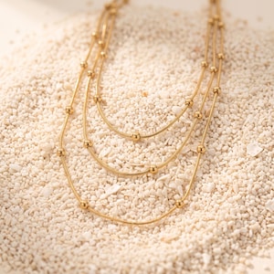 May include: A multi-strand gold-toned necklace featuring three delicate snake chains of varying lengths. Each chain is accented with small, evenly spaced round gold beads. The necklace is arranged in a layered, cascading design.