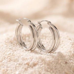 May include: A pair of silver-toned hoop earrings, each featuring a triple-layered design with three concentric circular bands. The earrings have a polished metallic finish and include a hinged post mechanism for secure closure at the top.