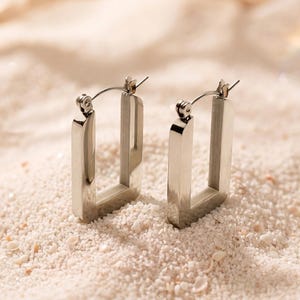May include: A pair of silver-toned hoop earrings featuring a rectangular, open-frame design with a polished metallic finish. Each earring includes a thin, hinged post mechanism at the top for secure closure, showcasing a clean and geometric aesthetic.