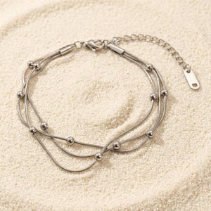May include: A silver-toned multi-strand bracelet featuring three delicate snake chains. The chains are adorned with small, evenly spaced round metal beads. The bracelet includes a lobster claw clasp and an adjustable extender chain with a small tag.