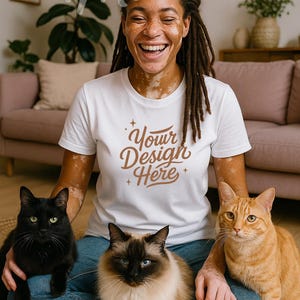 May include: A person wearing a white t-shirt with the text "Your Design Here" in brown script, sitting with three cats: a black cat, a Siamese cat, and an orange tabby. The person is smiling. The background includes a pink sofa and plants.