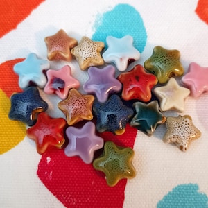 May include: A selection of star-shaped ceramic beads in a range of colours, such as blue, pink, red, and brown. The beads have a glossy finish and are displayed on a patterned fabric. These beads are ideal for jewellery making and craft projects.