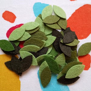 May include: A pile of felt leaf shapes in shades of green and brown, arranged on a white patterned fabric. The leaves are different sizes and textures, giving a natural look. Suitable for crafting and DIY projects.