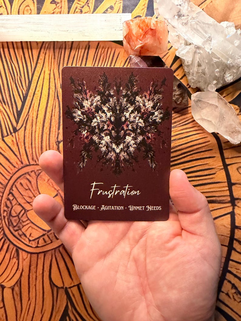 Pu&ograve; includere: Una carta dei tarocchi color amaranto con la parola "Frustration" in corsivo. La carta presenta un design astratto simmetrico in bianco e rosso scuro. Sotto, le parole "BLOCKAGE - AGITATION - UNMET NEEDS".