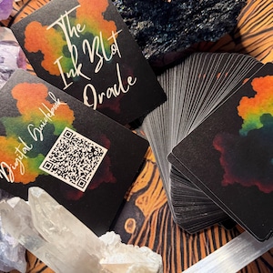 Pu&ograve; includere: Un mazzo di carte oracolo nere con un design a macchia di inchiostro colorato, intitolato "The Ink Blot Oracle". L'immagine include una guida con un codice QR, cristalli e altri oggetti spirituali. Le carte sono a ventaglio, mostrando i loro bordi.