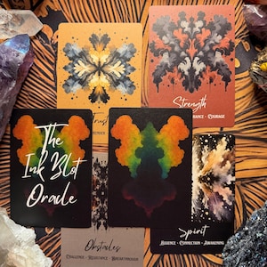 Pu&ograve; includere: Un set di carte oracolo con disegni astratti a macchie di inchiostro nei colori arancione, nero e arcobaleno. Le carte hanno titoli come "Forza" e "Spirito". Il mazzo si chiama "The Ink Blot Oracle". Cristalli e un tappetino fantasia circondano le carte.