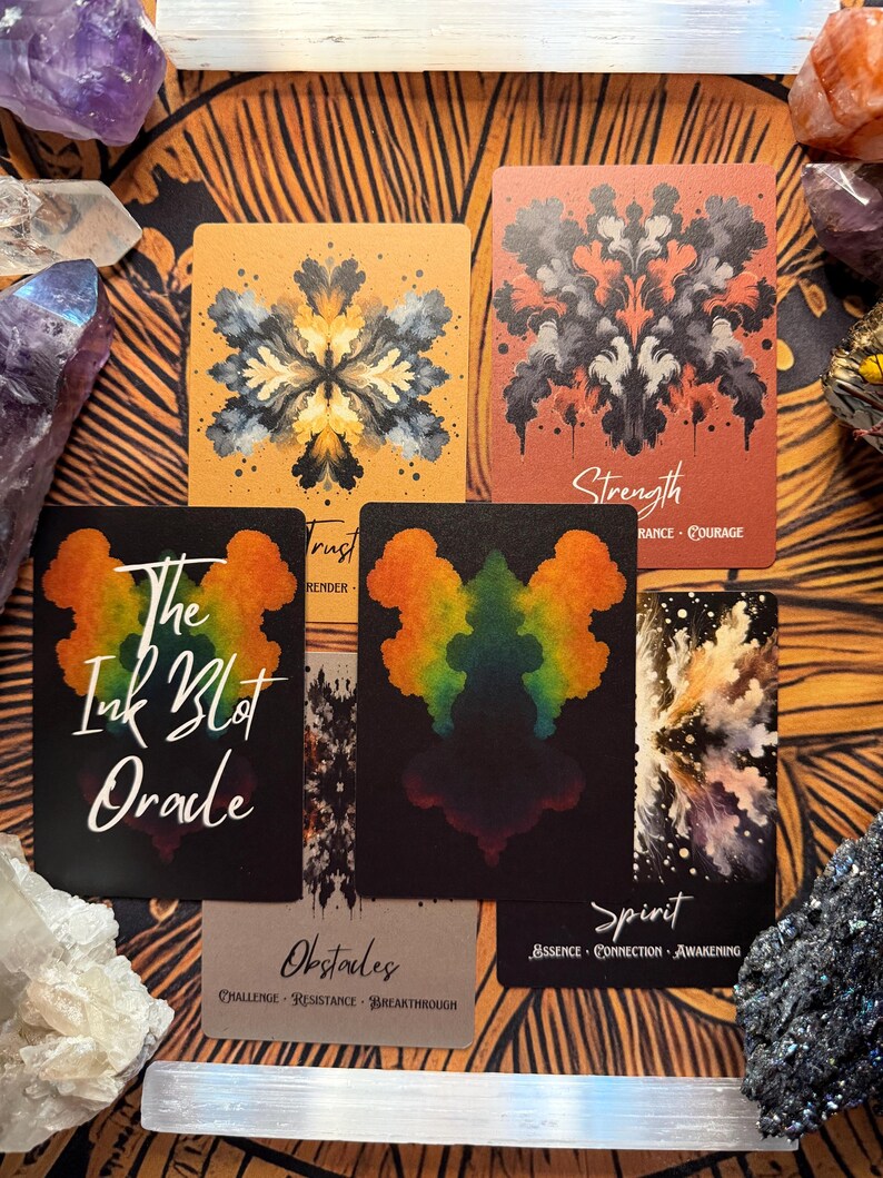 Pu&ograve; includere: Un set di carte oracolo con disegni astratti a macchie di inchiostro nei colori arancione, nero e arcobaleno. Le carte hanno titoli come "Forza" e "Spirito". Il mazzo si chiama "The Ink Blot Oracle". Cristalli e un tappetino fantasia circondano le carte.