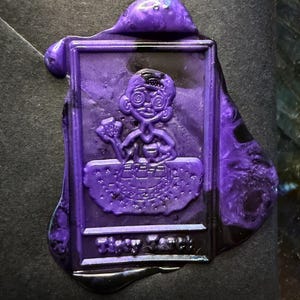 Pu&ograve; includere: Un sigillo di cera viola con un personaggio dei cartoni animati che tiene in mano un piccolo oggetto. Il sigillo ha una forma rettangolare con bordi arrotondati e la scritta "Tiny Cook" in basso. La cera ha una finitura lucida.