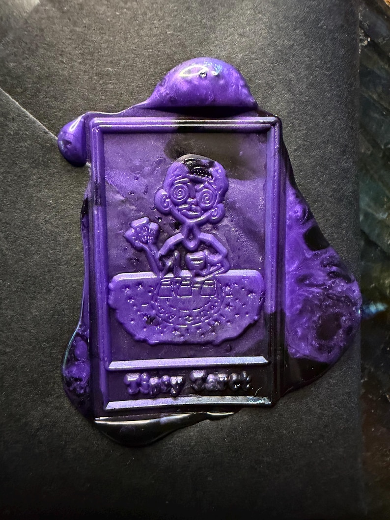 Pu&ograve; includere: Un sigillo di cera viola con un personaggio dei cartoni animati che tiene in mano un piccolo oggetto. Il sigillo ha una forma rettangolare con bordi arrotondati e la scritta "Tiny Cook" in basso. La cera ha una finitura lucida.