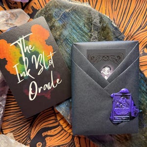 Pu&ograve; includere: Due mazzi di carte, uno con il testo "The Ink Blot Oracle" e un design a macchia di inchiostro colorato. L'altro mazzo &egrave; avvolto in carta nera e sigillato con un sigillo di cera viola. Lo sfondo include cristalli e una superficie a motivi.