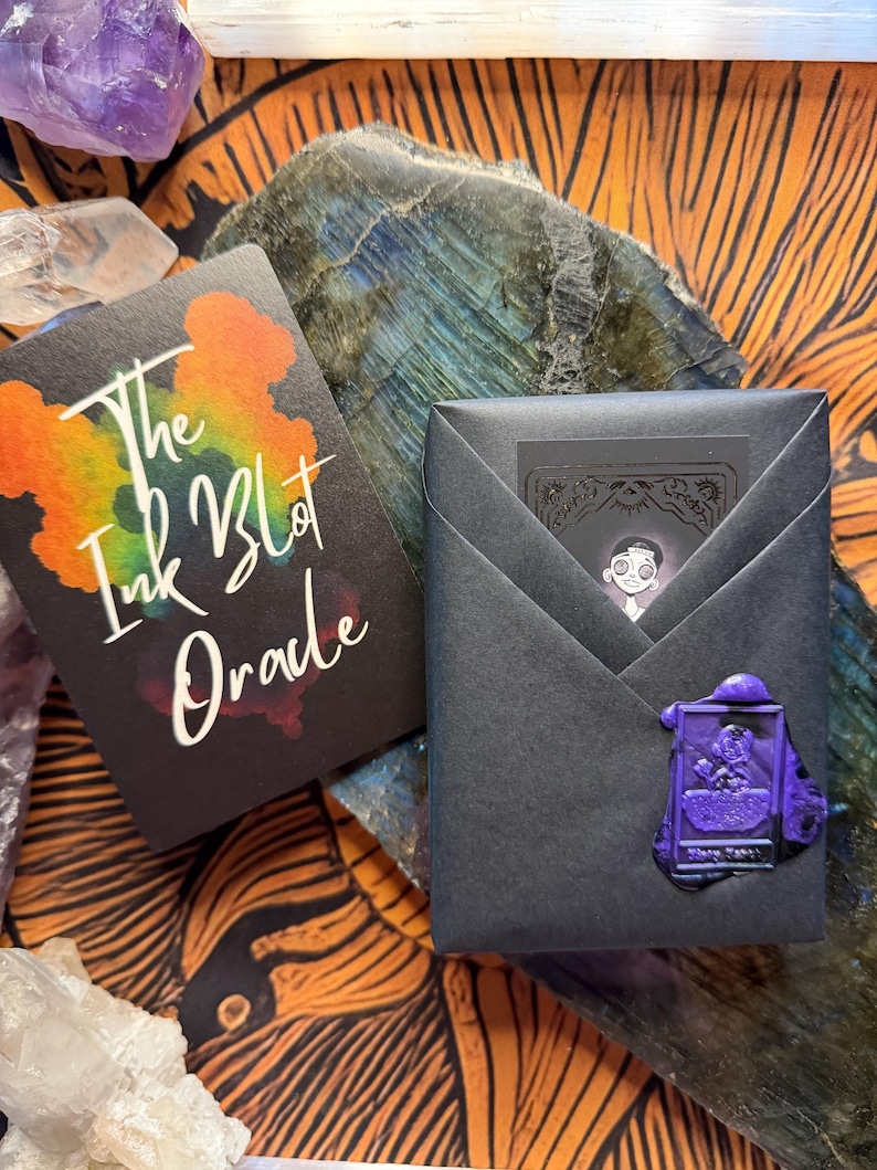 Pu&ograve; includere: Due mazzi di carte, uno con il testo "The Ink Blot Oracle" e un design a macchia di inchiostro colorato. L'altro mazzo &egrave; avvolto in carta nera e sigillato con un sigillo di cera viola. Lo sfondo include cristalli e una superficie a motivi.
