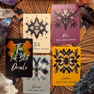 Pu&ograve; includere: Una vista dall'alto delle carte Oracle The Ink Blot con cristalli. Le carte presentano disegni astratti di macchie di inchiostro in nero, viola e giallo. Il testo sulle carte include "Truth", "Pain", "Cleanse" e "Caution". Cristalli di ametista, selenite e quarzo circondano le carte.