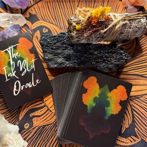Pu&ograve; includere: Un mazzo di carte oracolo nere con disegni colorati di macchie di inchiostro e la scritta "The Ink Blot Oracle" &egrave; appoggiato su una superficie a motivi arancioni e marroni. Sono presenti anche cristalli, un mazzetto di erbe essiccate e una pietra nera.