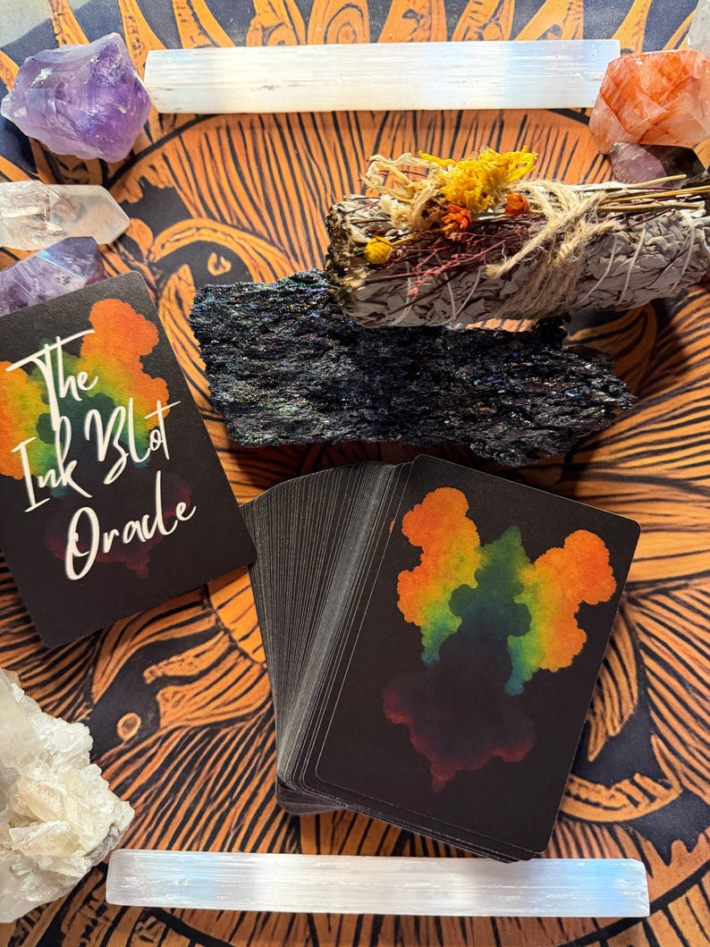 Pu&ograve; includere: Un mazzo di carte oracolo nere con disegni colorati di macchie di inchiostro e la scritta "The Ink Blot Oracle" &egrave; appoggiato su una superficie a motivi arancioni e marroni. Sono presenti anche cristalli, un mazzetto di erbe essiccate e una pietra nera.